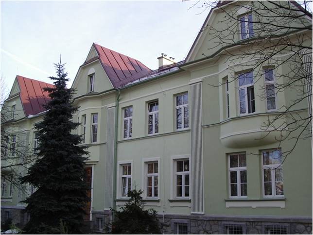 Widoczny jest zielono kremowy budynek z czerwonym dachem i wieloma oknami (białe ramy okienne). Widok budynku od strony ul. Żywieckiej.